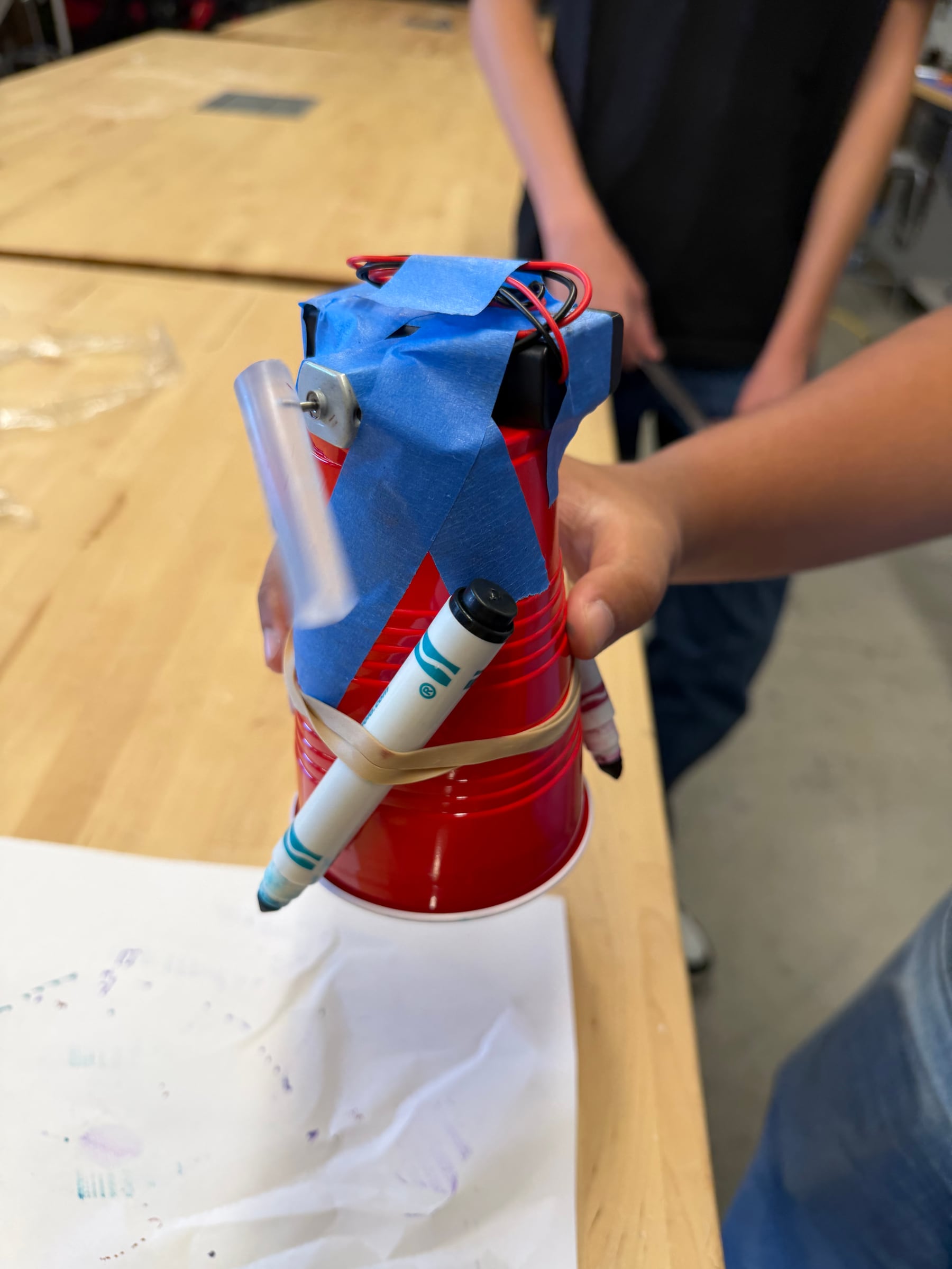 Student-built vibrating art robot with a cup body, marker legs, motor, and half glue-stick weight